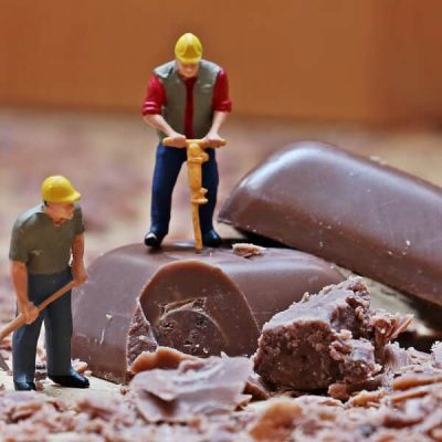 2 Trabajadores de plástico trabajando con pala y taladradora sobre una pastilla de chocolate de gran tamaño en comparación. Simboliza el trabajo mano a mano de consultoría de marketing online